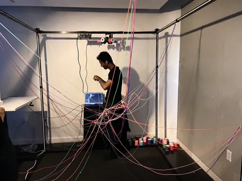 Leffin inside the metal frame placing threads while a participant converses at the desk
